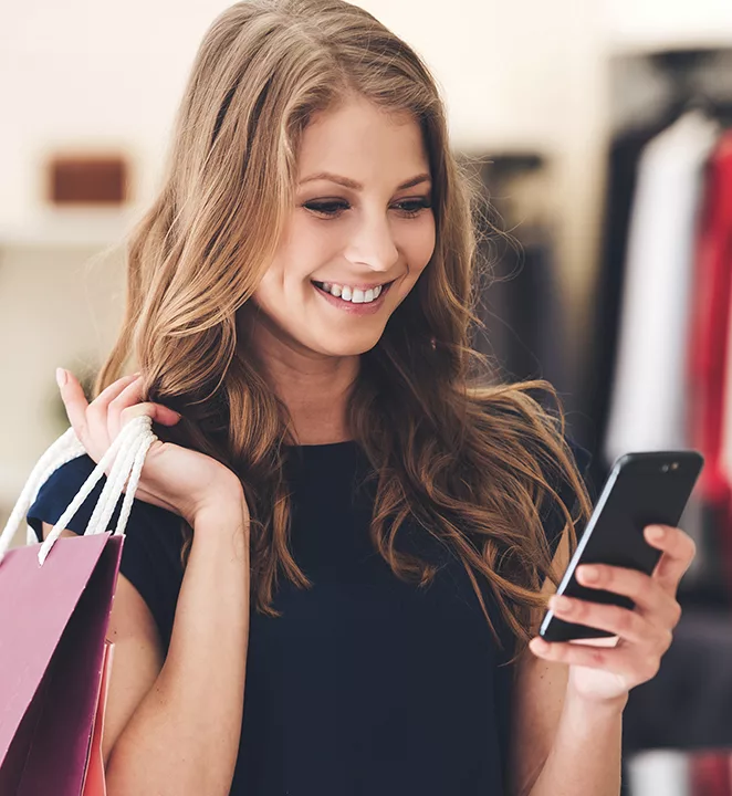 Woman smiling at her phone with a shopping bag in her hand. 